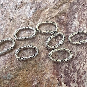 May include: Eight silver oval-shaped jump rings with a textured surface. The rings are open and have a slightly curved shape.