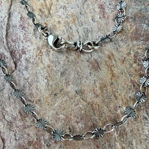 May include: A silver-toned metal chain necklace featuring a series of small, circular, sunburst-patterned discs linked together by oval rings. The necklace includes a secure lobster claw clasp mechanism for fastening the chain securely.