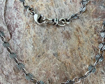 Può includere: Una collana a catena in metallo color argento caratterizzata da una serie di piccoli dischi circolari con motivo a raggiera collegati da anelli ovali. La collana include una chiusura a moschettone per fissare la catena.