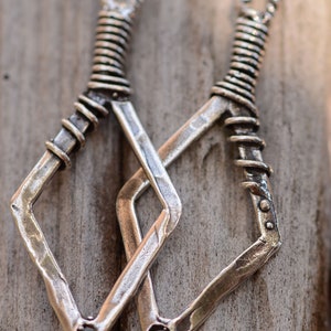 May include: Two silver-toned metal earrings with a diamond shape and a twisted wire design. The earrings are hanging from a wooden surface.