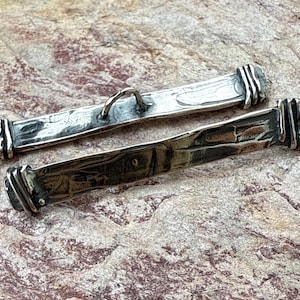 May include: Two silver bar clasps with textured surfaces and decorative ends. One clasp has a small loop. The clasps are displayed on a textured, neutral-toned surface. These clasps are suitable for jewelry making.