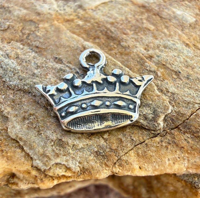 Puede incluir: Un dije de metal plateado con forma de corona que incluye una peque&ntilde;a anilla circular en la parte superior para su sujeci&oacute;n. La corona presenta una banda con formas de rombos en relieve y una serie de puntas redondeadas en el borde superior.