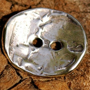 May include: A silver button with a textured design and two holes. The button is round and has a slightly raised edge.