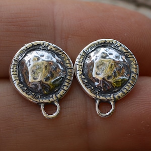May include: Two silver-toned metal pendant charms with a textured, hammered design. Each charm has a round, circular shape with a raised, textured center. The charms have a loop at the bottom for attaching to a necklace or bracelet.
