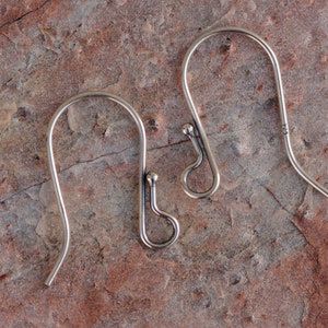 May include: Two silver earring hooks, each with a small ball at the top and a loop at the bottom. The hooks are on a brown, textured surface.