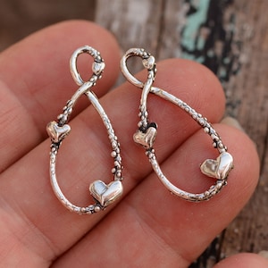 May include: Two silver-toned teardrop-shaped charms with heart accents. Each charm features a loop at the top and a heart at the bottom, with a textured, dotted design along the sides. The charms are displayed against a blurred wooden background.