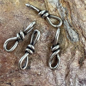 May include: Four silver-colored metal clasps, each featuring a knot design. The clasps are arranged on a textured, brown surface. The clasps are likely for jewelry or other craft projects.