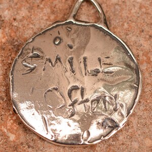 May include: Silver charm with a round, textured surface. The charm has a loop at the top for attaching to a necklace or bracelet. The words "Smile Often" are engraved on the charm.