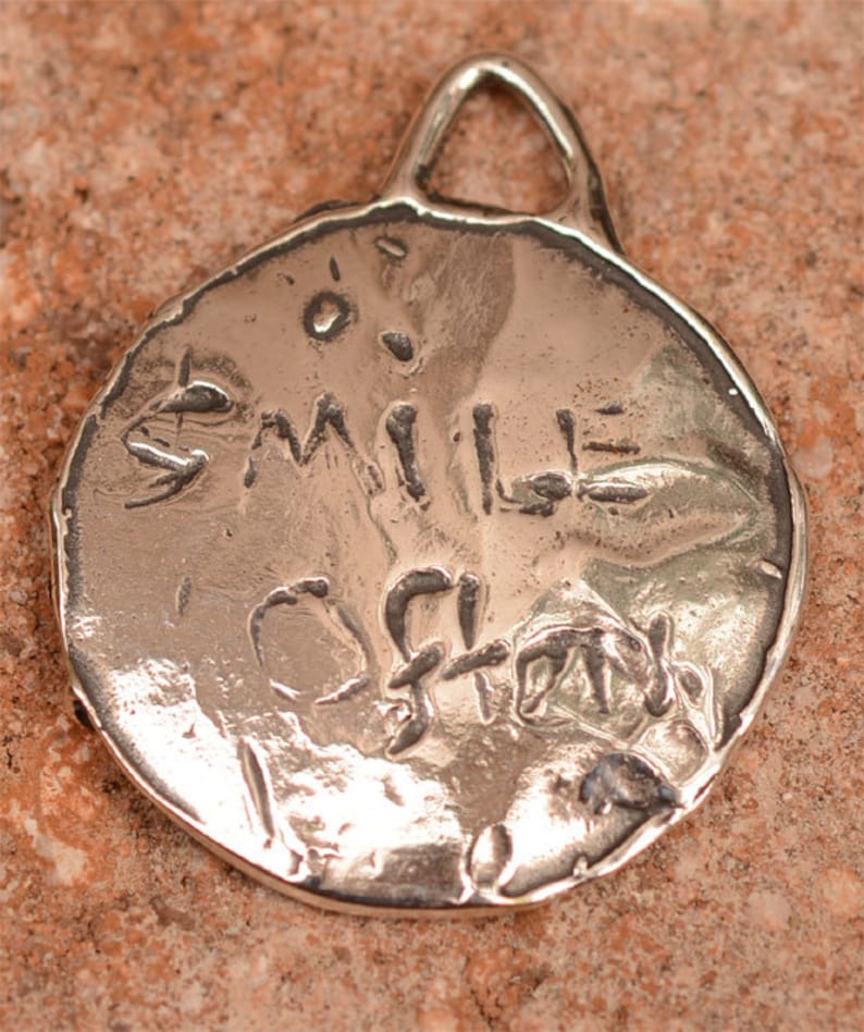May include: Silver charm with a round, textured surface. The charm has a loop at the top for attaching to a necklace or bracelet. The words "Smile Often" are engraved on the charm.