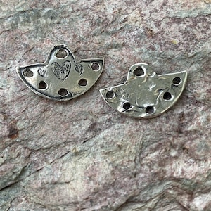 May include: Two silver metal charms in the shape of a half-circle with a heart design and holes around the edge.