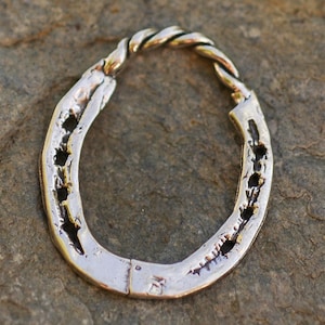 May include: A silver horseshoe-shaped pendant with a twisted rope design at the top. The horseshoe has several holes and a textured surface. The pendant is displayed on a stone surface.