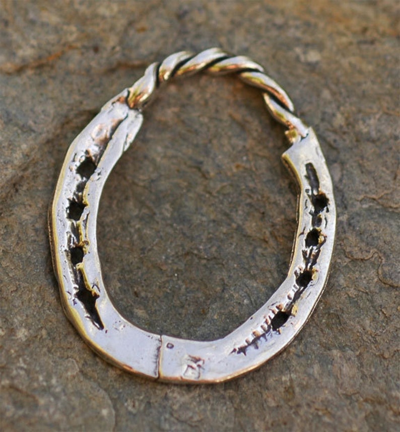 May include: A silver horseshoe-shaped pendant with a twisted rope design at the top. The horseshoe has several holes and a textured surface. The pendant is displayed on a stone surface.
