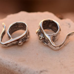 May include: Two silver-toned metal pendant bails with a textured finish and a loop on one end. The bails have a unique, organic shape and are designed to be used for attaching pendants to necklaces or bracelets.
