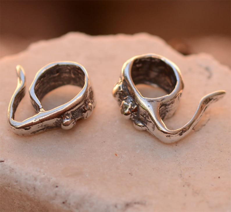 May include: Two silver-toned metal pendant bails with a textured finish and a loop on one end. The bails have a unique, organic shape and are designed to be used for attaching pendants to necklaces or bracelets.