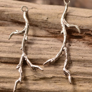 May include: Two silver metal twig-shaped charms with a loop at the top for hanging. The charms are detailed with textured branches and are perfect for jewelry making or crafting.