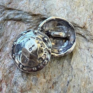 May include: Two silver-colored, hammered metal buttons with a textured surface and a raised rim. One button is face up, displaying the intricate details, while the other is face down, revealing the shank. The buttons are set against a neutral stone background.