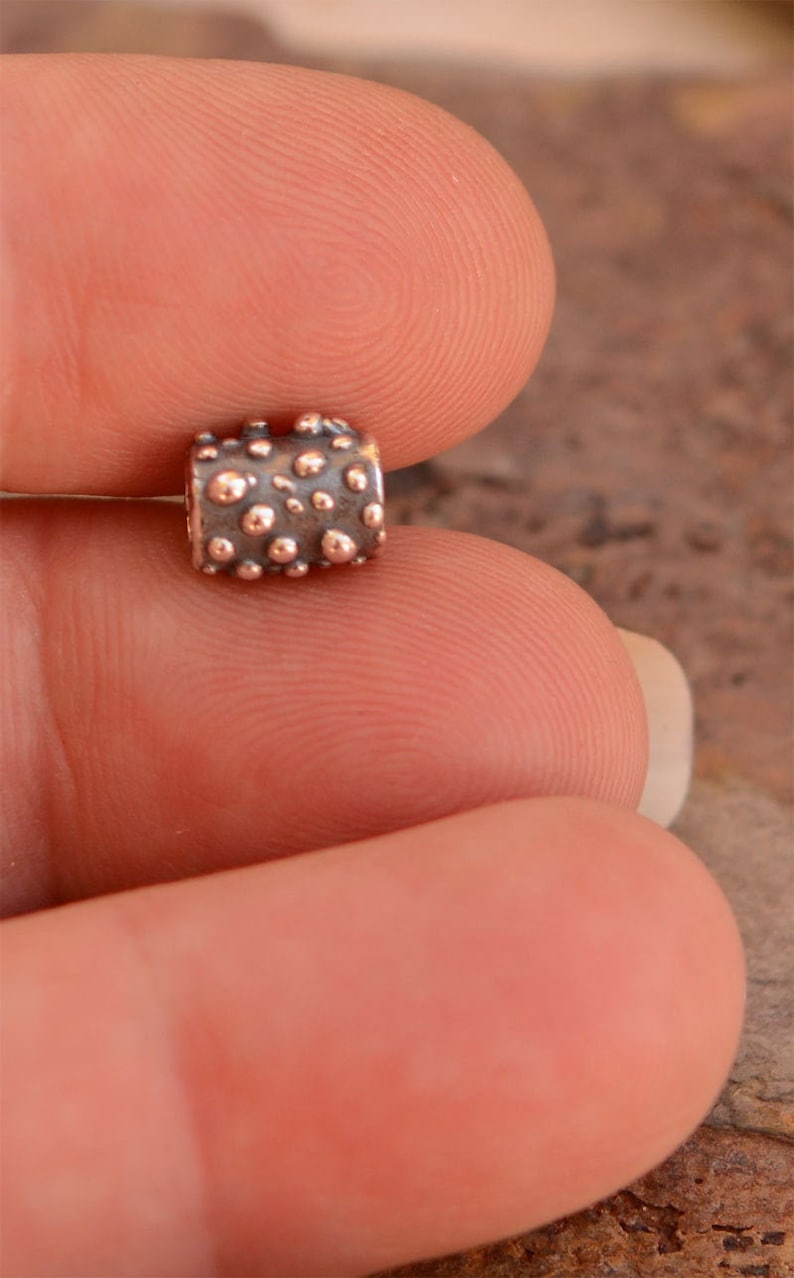 May include: A small, cylindrical bead with a textured surface. The bead is a dark gray color with raised, copper-colored dots. The bead is held between two fingers, showcasing its size and detail. This bead is likely for jewelry making.