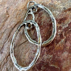 May include: Two interlocking oval-shaped pendants crafted from polished silver-toned metal. Each pendant features a textured, uneven surface with a hammered appearance. The two loops are connected at the top, creating a continuous, organic shape.