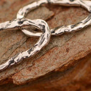 May include: Two silver rings linked together, with a textured, hammered finish. The rings are lying on a rough, brown surface.