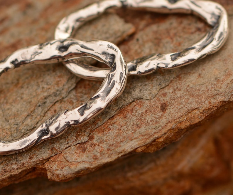 May include: Two silver rings linked together, with a textured, hammered finish. The rings are lying on a rough, brown surface.