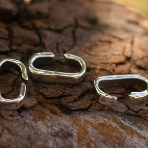 May include: Three silver metal jump rings, also known as connector rings, are shown on a brown wooden surface. The rings are oval shaped and have a smooth, polished finish.