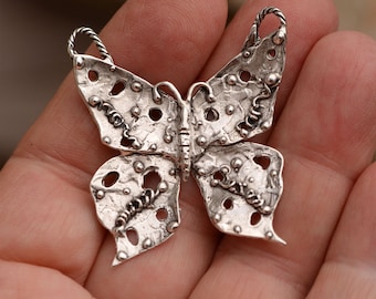 May include: A silver butterfly pendant with a textured, hammered finish and intricate cutouts. The pendant has a loop at the top for attaching to a chain.