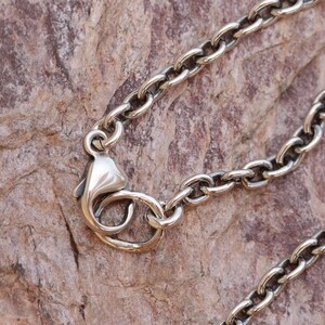 May include: Close-up of a silver-colored chain necklace with an oval link design. The necklace features a lobster clasp closure. The background is a textured, brown surface.