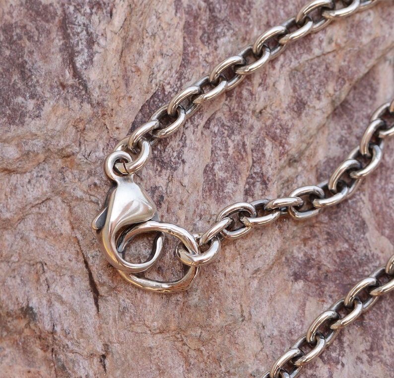 May include: Close-up of a silver-colored chain necklace with an oval link design. The necklace features a lobster clasp closure. The background is a textured, brown surface.