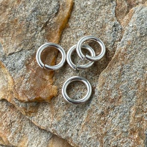 May include: Five silver jump rings, a type of jewelry finding, are arranged on a rough, brown, and tan rock surface.