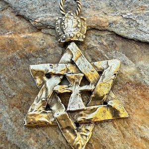 May include: A gold-coloured pendant featuring a Star of David design with a textured surface. The pendant includes a crown detail at the top and a cross-like symbol within the star. The pendant hangs from a twisted loop.