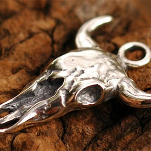 May include: A silver bull skull pendant with a loop for attaching a chain. The skull is detailed with textured bone and horns.