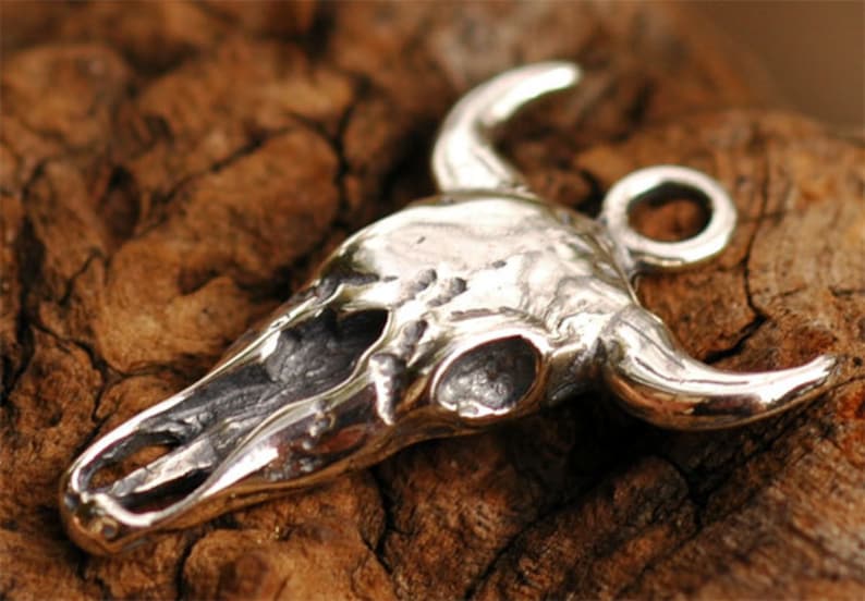 May include: A silver bull skull pendant with a loop for attaching a chain. The skull is detailed with textured bone and horns.