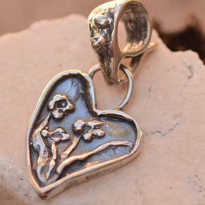 May include: A silver heart-shaped pendant with a floral design. The pendant has a loop at the top for attaching to a chain or cord.