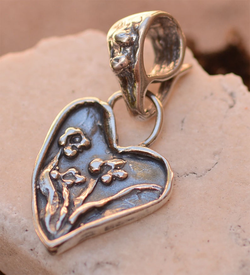 May include: A silver heart-shaped pendant with a floral design. The pendant has a loop at the top for attaching to a chain or cord.