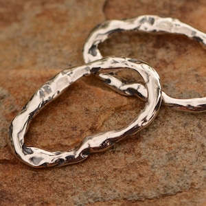 May include: Two silver oval-shaped rings with a textured, hammered finish. The rings are linked together and are lying on a brown, textured surface.