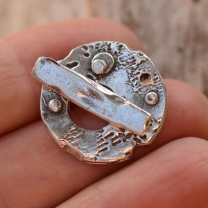 May include: A silver pendant with a textured circular design and a small, textured bar attached to the side.