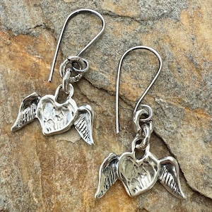 May include: A pair of silver-toned earrings. Each earring features a heart-shaped charm with detailed wings. The earrings have a hook design for easy wear. The background is a textured, neutral-toned surface.