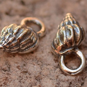 May include: Two silver-toned seashell charms with a loop for attaching to jewelry or other crafts. The charms are detailed with ridges and a textured surface.
