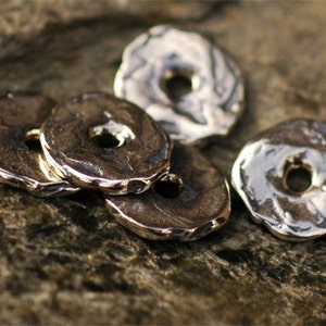 May include: Close-up of several silver-toned, donut-shaped beads with a textured surface and a hole in the center. The beads are arranged on a rough, brown stone surface, creating a rustic aesthetic.