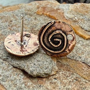 May include: A pair of copper-toned stud earrings. One earring features a spiral design, while the other shows the post and back. The earrings are round and have a textured surface. The earrings are displayed on a rough, stone surface.