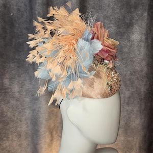 May include: A peach-colored fascinator with a decorative arrangement of feathers and fabric flowers. The headpiece features light blue and peach feathers, with a net veil and jewel accents. The base is a satin-like material.