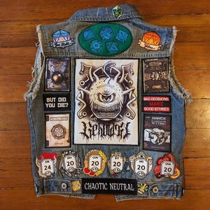 May include: A blue denim vest with various patches, including a large black and white patch with the word "Tendrils" and a smaller patch with the text "Bad Decisions Make Good Stories". The vest also has several dice patches, a patch with the text "But Did You Die?" and a patch with the text "CHAOTIC NEUTRAL".
