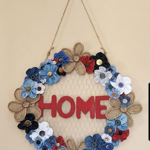 Könnte beinhalten: Ein kreisförmiger Wandbehang mit einem Hühnerdraht-Mittelteil, auf dem das Wort HOME aus rotem Stoff angebracht ist. Der Rahmen ist mit verschiedenen Stoffblumen in Blau, Rot und Weiß sowie Juteblumen und kleinen Knöpfen verziert.