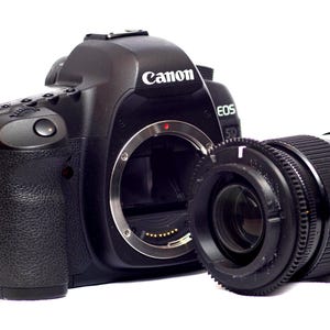 May include: A black Canon EOS 5D camera with a black lens attached. The camera has a silver ring around the lens and the word "Canon" printed on the top.