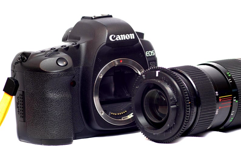 May include: A black Canon EOS 5D camera with a black lens attached. The camera has a silver ring around the lens and the word "Canon" printed on the top.
