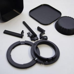 May include: A set of black plastic camera lens parts, including a lens hood, a lens cap, a gear ring, and a lens adapter ring. There are also several small black plastic pieces with letters on them.