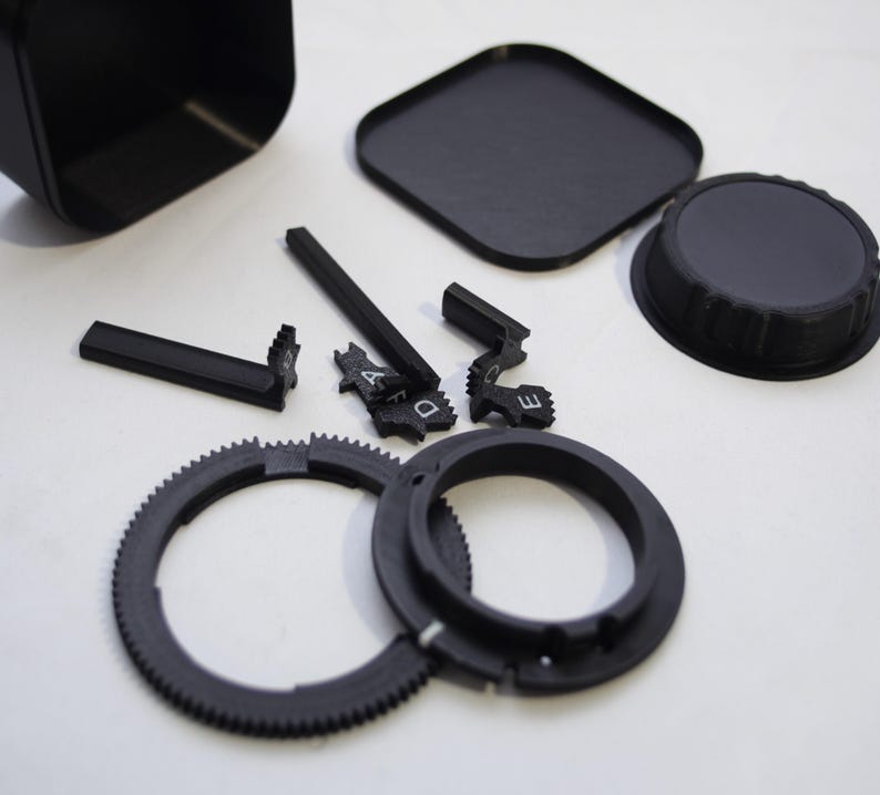 May include: A set of black plastic camera lens parts, including a lens hood, a lens cap, a gear ring, and a lens adapter ring. There are also several small black plastic pieces with letters on them.