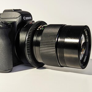 May include: A black Canon camera with a black Canon 135mm f/2.8 lens attached. The lens has a silver band with the text "Canon 135mm 1:2.8" printed on it.