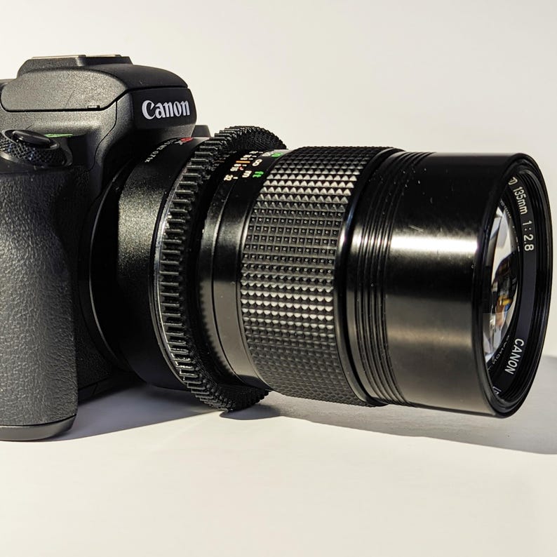 May include: A black Canon camera with a black Canon 135mm f/2.8 lens attached. The lens has a silver band with the text "Canon 135mm 1:2.8" printed on it.