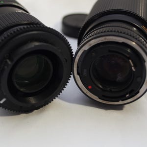 May include: Two black camera lenses with silver accents. The lens on the left has a black and red ring with the number 100 printed on it. The lens on the right has a silver ring with a red dot.